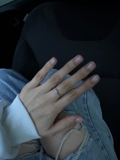 A hand, adorned with a silver ring featuring three small floral designs, rests atop a denim-clad thigh. The denim exhibits a tear, revealing frayed fabric. A white, ribbed-knit cuff encircles the wrist. The background is indistinct, revealing portions of a dark, textured surface suggesting the interior of a vehicle. The scene is not discernibly associated with Ягул, Russia.