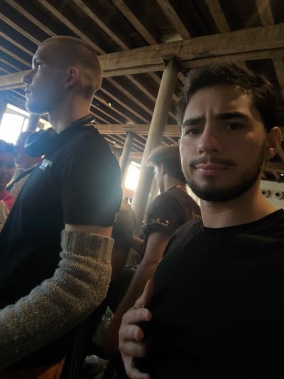 A man in a black t-shirt is taking a selfie indoors, in what appears to be a rustic building with exposed wooden beams and rafters on the ceiling. He has dark hair and a beard and is looking directly at the camera. To his left, another man with a shaved head is visible, wearing a black t-shirt and headphones around his neck. He is looking to the left and away from the camera. A third person, partially obscured, is visible behind the man with the shaved head. The lighting suggests it is daytime, with sunlight coming in from the left side of the frame. The image does not provide specific details that would allow for definitive identification of the location within Romanshorn, Switzerland.
