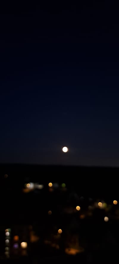 The image captures a nighttime cityscape, likely taken from an elevated position, overlooking Zhukovo, Russia. The sky is a deep, dark blue, with a bright, full moon positioned slightly off-center in the upper portion of the frame. Below the sky, a dark, blurred horizon line suggests the silhouette of buildings and landforms. The foreground is dominated by out-of-focus lights, appearing as bokeh orbs of varying colors, primarily yellow and white, scattered across the scene, indicating the presence of streetlights and illuminated buildings. The overall impression is that of a quiet, nocturnal urban environment illuminated by the moon and artificial lights.