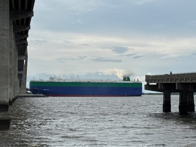 The image shows a large car carrier ship, the "CRYSTAL BAY," sailing on water. The ship is predominantly blue and green with a red waterline. The ship is viewed from under a bridge, the side of which is visible on the left of the frame. The water is choppy and grey. On the right side of the image, another bridge structure with concrete pillars is visible, and a person is standing on it. The sky is overcast with some clouds. The scene appears to be in an industrial port area, potentially in the city of Brunswick, Georgia, United States of America, known for its automobile import/export facilities.