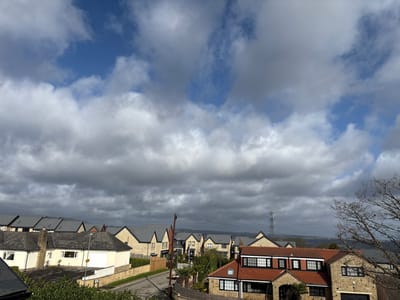 This image depicts an elevated view of a residential area in Kirklees, United Kingdom, under a partly cloudy sky. The upper portion of the frame is filled with extensive, dense grey and white stratocumulus clouds, with visible patches of clear blue sky in the top-left and top-right sections.
In the foreground, a dark grey roofline is visible at the bottom left, alongside green foliage. To the bottom right, bare, dark tree branches extend into the frame. The immediate middle ground features a light-colored detached bungalow with a dark grey tiled roof, a white garage door, and light-colored perimeter wall, situated beside a paved residential street. A dark utility pole with overhead lines stands prominently in the mid-left area.
The scene extends into the mid-ground and background, showcasing numerous residential properties of varying architectural styles. These include stone-clad houses with distinct red-tiled roofs and multiple black-framed windows, as well as houses with light-colored rendered facades and dark grey tiled roofs. The landscape slopes downwards and then rises in the distance, populated by further clusters of houses. A large metal electricity transmission tower (pylon) is discernible on a distant elevated ridge or hill. The far horizon shows a hazy, undulating landscape beneath the cloud cover. No individuals or dynamic interactions are observable within the scene.