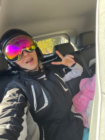 A person is depicted seated in the rear passenger seat of a vehicle. The individual, positioned slightly off-center to the left, is wearing a black ski helmet adorned with large, highly reflective, multi-chromatic ski goggles that display prominent pink, orange, yellow, and blue hues. The lower portion of the face is visible, with the mouth slightly open. The person is dressed in a black ski jacket featuring white and light grey panels and a white vertical zipper on the chest. The right hand is raised, displaying a "peace" sign with the index and middle fingers extended; a silver-colored ring is visible on the ring finger. The vehicle's interior shows black upholstered headrests and seatbacks, along with a light-colored ceiling. Through the rear side window, blurred green foliage and bright outdoor light are visible, indicating an exterior environment. On the seat to the right of the person, a crumpled or folded pink garment is visible amidst other light-colored fabric items. The specific street or address within Dulovine, Montenegro, cannot be determined from the visual information provided.