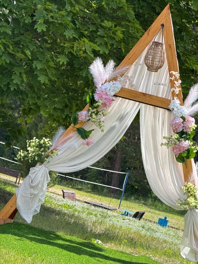An outdoor scene in Yagodnoe, Russia, depicts a decorated triangular wooden archway under bright daylight. The arch, constructed from light brown timber, features a flat horizontal crossbar connecting the two upper sides. White, translucent fabric is draped and tied around the arch's structure, creating folds and swags. Integrated into the fabric drapes and wooden frame are floral arrangements consisting of pink hydrangeas, white small flowers, and green foliage. Several long, light pink and white feathers are also incorporated into these decorative clusters. A single woven wicker lantern hangs from the apex of the arch. Small white lights are visible along the underside of the top crossbar.
The background primarily consists of dense green foliage from deciduous trees, indicating a natural outdoor environment. In the midground, behind the arch, an area of lighter green grass and sparse vegetation is visible. A blue-framed volleyball net and pole are present in the mid-distance, slightly to the left of the arch's center. Scattered dark-colored objects, resembling benches or equipment, are visible on the grass in the midground. The immediate foreground features a patch of vibrant green, short-cut grass or artificial turf with distinct, elongated shadows cast upon it, suggesting strong sunlight. No individuals are present in the scene.