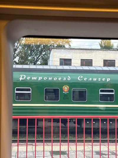 A side view of a green train car with white Cyrillic lettering reads "Ретропоезд Селигер" (Retro-train Seliger). A circular emblem with a crest is located to the left of the lettering. Below the emblem, the numbers "006 32190" are visible. The train car has several rectangular windows with white frames. In the foreground, a red metal railing is in focus, with train tracks and a platform visible behind it. The train is parked at a station, likely in Bologoe, Russia, given the "Seliger" designation, which refers to Lake Seliger, a popular tourist destination near the city. The background shows a brick building with windows. The overall scene suggests a train station environment.