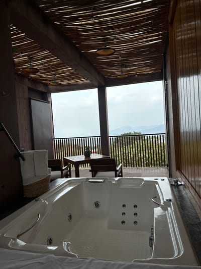 This image depicts an indoor/outdoor spa area in Tepoztlán, Mexico. The right side is dominated by a white jacuzzi tub, oriented vertically in the frame. To the left of the tub is an outdoor area with a wooden fence overlooking a mountainous landscape under a cloudy sky. A wooden table and chairs sit on the patio. The spa area's ceiling features a decorative design made of wood or bamboo slats, punctuated by light fixtures. Underneath the outdoor section, folded white towels and a decorative cushion are placed. A black railing is partially visible, leading up to the spa area. The overall impression is of a luxury spa facility with a view of the Tepoztlán landscape.