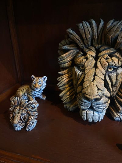 A close-up shot presents two ornate, tiger-themed carvings against a dark, polished wooden surface. The larger carving, positioned above, depicts a tiger's head, sculpted with stylized fur and piercing eyes. Below, a smaller, full-bodied tiger carving is angled as if climbing. The carvings exhibit a rough, textured finish with a color palette of bronzes, grays, and creams, enhancing their three-dimensional appearance. The wooden surface provides a stark, dark backdrop, emphasizing the carvings' intricate details and highlighting the interplay of light and shadow. There is no evidence of location.