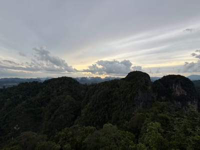A panoramic, high-angle, outdoor shot captures a dense, dark green forest covering rolling hills and jagged, rocky outcrops under a cloudy, overcast sky. The horizon is lined with distant, hazy mountain ranges. The lighting suggests either early morning or late afternoon. The scene is devoid of any people or structures. The location is identified as being in Ban Khao Tang, Thailand.