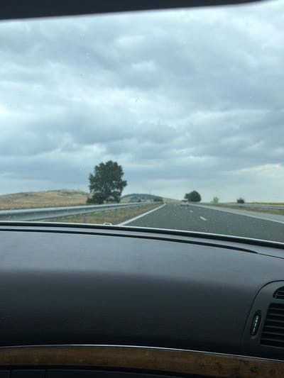 The image shows a view from the interior of a vehicle looking out through the windshield. The windshield has water droplets on it. The scene outside is a highway with a cloudy, overcast sky. There are fields on either side of the road, with a solitary tree visible in the distance to the left. Other vehicles can be seen on the highway. The foreground of the image shows the dashboard of the car, with a textured surface and a strip of wood-grain trim. The location is identified as Polsko Padarevo, Bulgaria.