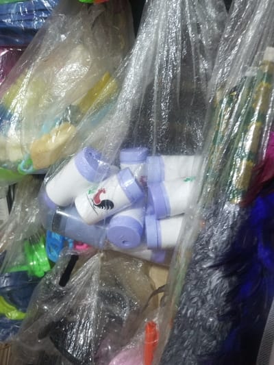 Various items in transparent plastic packaging fill the frame. Predominantly visible are stacks of white plastic containers with purple lids and rooster logos. Also noticeable are containers of green and yellow objects, and a cluster of blue and pink items towards the bottom. The shot appears to be inside a storage space or a store.