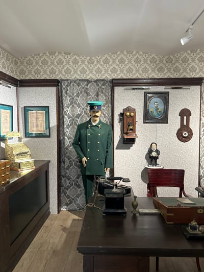 The image depicts a room furnished as a historical display, likely in Vyatskoye, Russia, recreating an old office or reception area. A mannequin dressed in a green military-style uniform with a cap stands prominently in the foreground. The room's walls are adorned with patterned wallpaper.

Various objects suggestive of a past era are visible. On the wall, there's an antique-looking thermometer, a framed oval portrait of a man in uniform, and an old wooden wall-mounted telephone with a receiver. A vintage desktop telephone with a handset is also present on a desk. A wooden chair with a dark red seat and back is positioned near a desk. The desk itself is dark wood and appears to be a central piece of furniture, holding several items including what looks like an inkwell and a pen holder. To the right of the desk, a vintage cash register is visible. Framed documents, possibly certificates or historical papers, are hung on the wall.

The scene appears to be indoors, with artificial lighting. The overall atmosphere suggests a museum exhibit or a meticulously preserved historical space. The objects and attire point towards a setting from the late 19th or early 20th century. There is no clear indication of the time of day or weather outside. The mannequin's pose is neutral, and there are no visible expressions on any depicted figures. Some text is visible on the cash register, including "Amount Purchased" and what appears to be a price or serial number.
