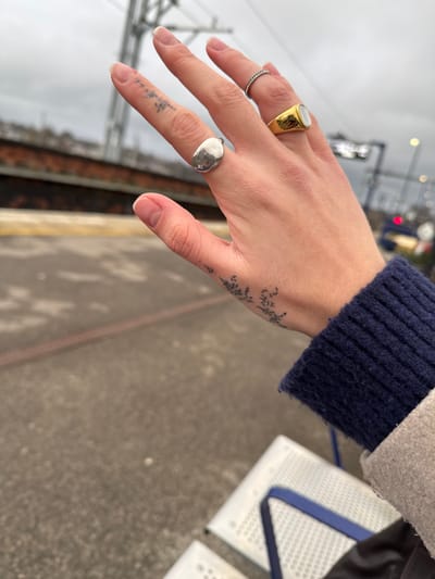 A pale hand adorned with three rings is in focus. A gold ring with a possible mother-of-pearl inset is on the ring finger, a thin silver band on the middle finger, and a silver signet ring on the index finger. Tattoos are visible on the thumb and along the side of the hand, near the wrist. A blue knit sleeve peeks out from a grey or beige coat. The hand is positioned over a grey platform with the edge of a train track visible in the background.