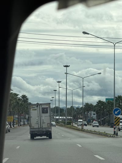 The image depicts a view from inside a vehicle, looking out at a road in Ban Ta Khun, Thailand. The sky is overcast with dramatic, voluminous clouds. A line of tall streetlights with multiple lamps stretches along the right side of the road, interspersed with palm trees. On the left, more palm trees line the road, with some buildings visible behind them.
The road is a multi-lane asphalt surface with white lane markings. A white box truck is prominently featured in the foreground, traveling in the same direction as the viewer. Its rear doors are closed, and a license plate with Thai characters is visible. Several other vehicles are on the road, including cars and a van. A blue directional sign with a white arrow pointing left is positioned on the right side of the road. Traffic appears to be moving at a moderate pace. The overall impression is of a somewhat busy, but not congested, road in a tropical or subtropical setting.