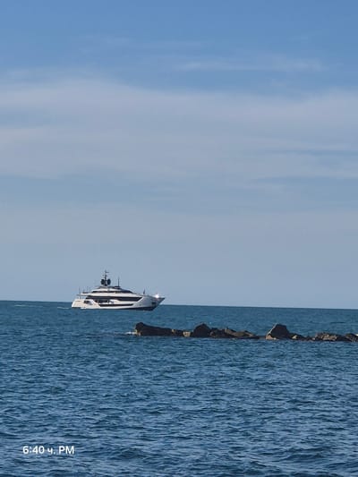 A large white yacht with black trim is sailing on a calm, blue sea. The yacht has multiple decks and appears to be luxurious. A line of dark, rocky breakwaters stretches across the water in the foreground, with some of the rocks emerging above the surface. The sky above is a clear, pale blue with some wispy white clouds. A timestamp in the lower left corner reads "6:40 PM". The image appears to have been taken from a shoreline or another boat. The scene is consistent with a view from the coast of Bisceglie, Italy, given the typical appearance of the Adriatic Sea and its coastline.