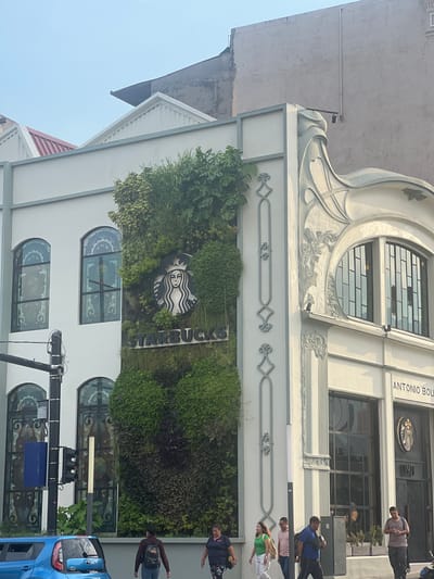 The image shows the exterior of a Starbucks store located in San Salvador, El Salvador. The building has a white facade with distinctive architectural features, including arched windows with decorative stained-glass, intricate molding details, and a vertical garden incorporating the Starbucks logo. Signage above the entrance indicates the address as "Antonio Boulevard." Several pedestrians are visible on the sidewalk in front of the building. A blue car is parked on the street to the right. A traffic signal is also present. The sky is clear and light blue.