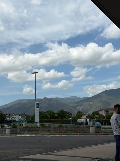 This image captures a bright daytime scene at a roadside location in Castrocielo, Italy, set against a dramatic mountainous backdrop. The photograph is oriented vertically (rotated 90 degrees clockwise from its likely original capture).
In the foreground, a paved road or shoulder occupies the lower right portion of the frame, bordered by a series of orange traffic cones. A person, visible from the chest up, stands on the far right, wearing a blue and white plaid shirt and blue jeans, looking towards the left side of the frame.
The mid-ground features a tall, grey light pole with multiple fixtures at its top, positioned centrally. To its right, a prominent white sign with a blue and orange logo is visible, bearing the text "Q8 Easy" and partially "Caldaie & Co", indicating a fuel station or service area. Below this, a green road sign provides directions, displaying an arrow pointing right and the Italian word "USCITA" (Exit), along with pictograms for services such as a car, truck, and possibly an electric vehicle charging point. Several buildings with light-colored walls and numerous trees and green foliage are scattered between the road and the distant mountains.
The background is dominated by imposing, rugged mountains covered in varying shades of green, suggesting a verdant landscape. The sky above is partially cloudy, with patches of clear blue interspersed with large, fluffy white clouds. The scene conveys an impression of a well-maintained service area amidst a scenic, natural environment.