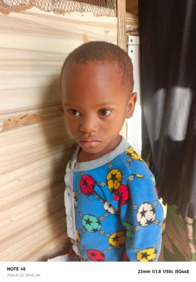 The image depicts a young child standing indoors in Panshekara, Nigeria. The child, with dark skin and short, dark hair, wears a blue long-sleeved shirt patterned with multiple interconnected soccer balls in red, yellow, green, and white/black. A contrasting light grey or white panel is visible on the garment's left shoulder and upper chest area. The child maintains a neutral expression, gazing slightly towards the viewer's right. To the child's left, a light-colored wooden wall or paneling shows some minor damage and exposed wire mesh near the top. To the child's right, a dark fabric, likely a curtain, is partially visible behind a white vertical structural element. The scene is consistent with an indoor domestic setting.