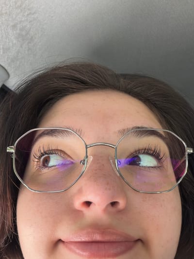 A close-up, low-angle shot shows a young woman wearing glasses and looking upwards. Her dark hair frames her face, and her eyes are visible through the hexagonal lenses of her glasses. The reflection on the lenses shows purple and pink hues, likely from lighting. Her skin has a scattering of light freckles across her nose and cheeks. Her lips are slightly parted. The background is a blurred, textured gray surface, possibly the interior of a vehicle. The image does not contain any elements that would allow for a definitive location within Nakhabino, Russia.