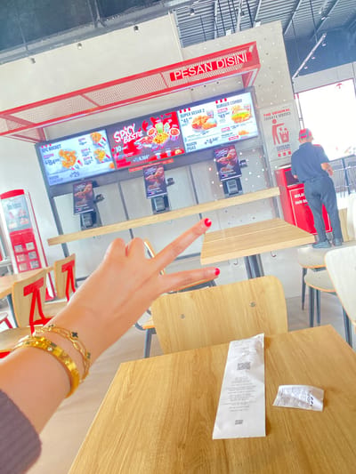 This media file shows a view from a table in a KFC restaurant, likely in Lhoksukon, Indonesia. The scene is an interior dining area with wooden tables and chairs. A person's hand, adorned with two gold bracelets, is in the foreground, making a peace sign gesture.
Behind the hand, digital menu boards are visible, displaying various KFC meal deals and prices in Indonesian currency (Rp). Items like "KFC BUCKET," "SPICY TE BESTIE," "BURGER COMBO," and "SUPER BESAR 2" are advertised, with prices ranging from Rp34,545 to Rp154,000. Some menu items are accompanied by images of fried chicken and burgers. The restaurant branding "KFC" and slogans such as "PESAN DISINI" (Order Here) and "MAKIN PUAS" (More Satisfied) are prominent.
In the background, a person in a dark blue uniform and a red cap is walking away from the camera. There are also receipt papers visible on one of the tables, with one small tag bearing the "mandırı" logo, possibly indicating a payment method. The lighting suggests it is daytime. The overall mood appears casual and possibly relates to ordering food or enjoying a meal.