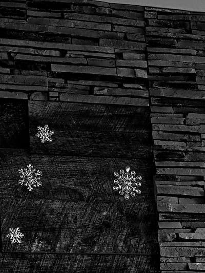 The scene, located in Donchevo, Bulgaria, depicts a section of an interior wall rendered in black and white. The wall surface is composed of two distinct materials: a dark, stacked stone veneer on the right side and upper portion, and dark, horizontally laid wooden planks on the left side and lower portion. The stone veneer consists of irregularly shaped, flat rectangular stones arranged in courses. The wooden planks feature visible grain and texture. Four metallic, snowflake-shaped decorative objects are affixed to the wooden planks. Three smaller, similarly sized snowflakes are positioned on the upper-left, mid-left, and lower-left areas of the wooden section. A fourth snowflake, larger and more intricately designed, is located on the mid-right of the wooden section, near the vertical interface with the stone veneer. No individuals, actions, or specific interactions are depicted.