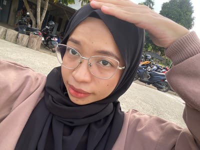 A young woman wearing a black hijab and a pink long-sleeved shirt is taking a selfie outdoors. She has glasses on and her left hand is raised to her forehead, partially obscuring the top of her head. The background shows several parked motorcycles on a paved surface. To the left, there are wooden posts and a building with trees and foliage. The overall scene appears to be in a casual outdoor setting, possibly a street or parking area. Based on visual cues like the architecture and parked vehicles, the location is identified as Air Molek, Indonesia.