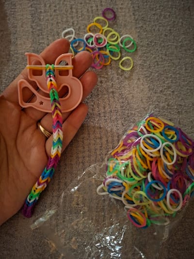 An indoor scene displays various items for creating rubber band crafts on a textured, neutral grey surface. A human hand, adorned with a gold-toned ring on the ring finger, holds a pink plastic loom tool. A partially completed multi-colored rubber band chain, exhibiting a braided pattern in vibrant hues such as purple, blue, green, yellow, orange, and white, extends downwards from the tool. Scattered across the surface near the hand are numerous loose rubber bands in a mix of colors including yellow, green, blue, purple, and white. To the lower right, a transparent plastic bag is partially visible, containing a dense collection of similarly multi-colored rubber bands. The specific location within the city of Davos, Switzerland, is not discernible from the visual content.