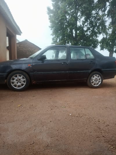 The image shows a dark-colored sedan parked on a dirt road in front of a building with a corrugated metal roof. The building appears to be made of earth or brick. To the right of the car are large trees with green foliage. The sky is overcast. There are no people clearly visible in the image. The car is a Volkswagen Golf with silver alloy wheels. The car appears to be older and shows some signs of wear and tear, with rust visible on the lower body panels. The ground is composed of reddish-brown dirt with scattered small stones and debris. This scene is consistent with a rural or semi-urban setting in Zot Shen, Nigeria.