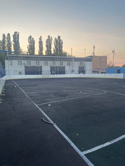 The image depicts an outdoor basketball court with black asphalt and white markings. A basketball hoop with a blue pole and red rim is visible on the left side. Adjacent to the court is a multi-story building with white and gray facades, featuring numerous windows and air conditioning units. Trees with dense foliage line the area between the court and the building. In the background, there are other buildings, including a light brown brick structure, and utility poles with wires. The sky is clear and light, suggesting it is either early morning or late afternoon. The scene appears to be set in a city or town, given the presence of buildings and infrastructure. There is no visible text in the image. The overall impression is one of an urban recreational space under a calm, possibly twilight, sky.