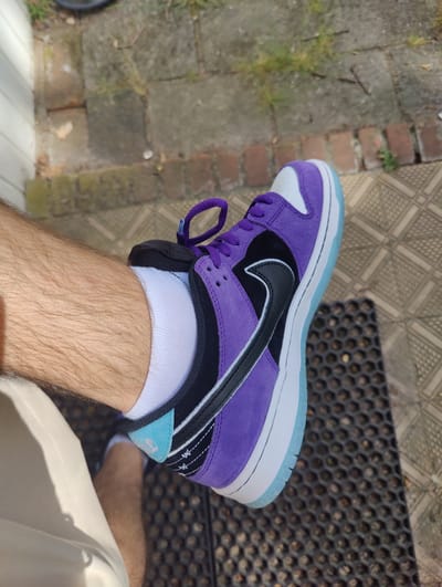 A person wearing a Nike Dunk Low SB purple and white sneaker is seen in an outdoor setting, likely in Dorst, Netherlands. The shoe is predominantly purple suede with a white toe box and a black Nike swoosh. The midsole is white with an icy blue outsole. The person is also wearing a white sock and khaki shorts. The background includes brick paving, a grey doormat, and a white wall.