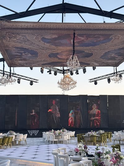 This image depicts an outdoor event setup in Khirbet Al Souq, Jordan. The scene is dominated by a large, ornate fabric ceiling that appears to be a tapestry, suspended from a dark metal framework. Below this canopy hang several elaborate crystal chandeliers and a series of stage lights.

The background features a black, multi-panelled screen or wall, onto which are projected large, stylized images of figures dressed in what appear to be traditional or theatrical costumes, rendered with a digital, pixelated effect.

In the foreground, a reception or dining area is arranged on a checkered white and grey tiled floor. Several round tables are set with white chairs and covered with floral centerpieces. The chairs are of varying styles, some upholstered in olive green and others in white. The overall impression is of a luxurious outdoor event venue. The sky above is clear and light blue, suggesting daytime or early evening.