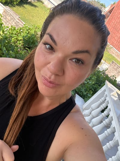 A selfie depicts a woman with fair skin, brown hair, and brown eyes, posing outdoors. She wears a black tank top. Behind her, there is green foliage and a white decorative railing, suggesting a balcony or porch. In the distance, a red-tiled roof can be seen. Based on the prompt, the location is Bijela, Montenegro.