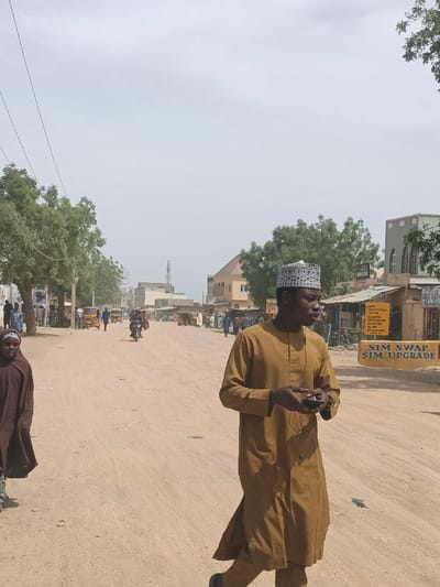 An overhead-level street scene in Hotoro North, Nigeria, depicts a dusty, unpaved road under an overcast sky. In the immediate foreground, a male individual walks towards the viewer's left. He wears a mustard-brown long-sleeved tunic, a white cap with dark geometric patterns, and dark shoes. He holds a dark-colored smartphone or device in his left hand with wired earphones around his neck and a watch on his left wrist. Partially visible on the far left, a female figure in a dark brown flowing garment and matching head covering walks in the same direction.
The midground features the wide, light brown dirt road with visible vehicle tracks. Multiple yellow auto-rickshaws (tricycles) are parked or moving along the road, alongside pedestrians. A motorcycle with a rider proceeds away from the camera in the distance.
On the left side of the road, a line of green-leafed trees precedes various low-rise buildings. On the right, several commercial structures are visible. A prominent sign on the right reads "SIM SWAP SIM UPGRADE" in yellow text on a dark background. Further back, buildings with varying facades, including a cream-colored building with a reddish-brown roof and a grey structure with arched windows, are present. Power lines span the upper frame, and a cellular tower is visible in the distant background. Numerous people are scattered throughout the scene, primarily pedestrians.