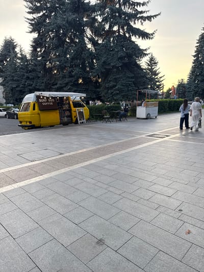 This media file depicts an outdoor scene in Khamkhi, Russia, during what appears to be either early morning or late afternoon due to the soft, diffused light. The image is captured with a vertical orientation, and the scene is dominated by a large, lush green coniferous tree on the left side.
In the foreground, a bright yellow van, labeled "SURF BUS COFFEE," is parked along a paved walkway. The van is open, revealing stocked shelves and possibly a service counter inside, suggesting it is a mobile coffee shop. The walkway is made of large, light grey rectangular tiles, bordered by a lighter strip.
Further back on the walkway, a smaller, white food cart with the text "Sweet Dreams" is visible. Two people are walking away from the camera on the walkway, one in a white outfit and another in a blue shirt and dark pants. Another person is seated at a table with chairs near the food cart, facing away from the camera.
The background shows more greenery, including bushes, and hints of buildings with red roofs. The sky is a pale yellow, indicating the time of day. The overall impression is one of a relaxed, public space with active small businesses catering to passersby.