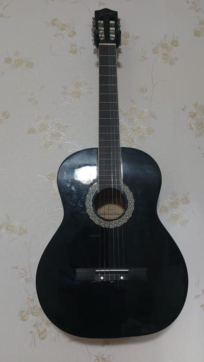 A black acoustic guitar is mounted on a white wall with a floral pattern. The guitar is positioned vertically, with its headstock at the top and its body below. The headstock is dark colored and features tuning pegs. The neck of the guitar is dark and has frets. The body of the guitar is black and glossy, reflecting the surrounding light. There is a soundhole in the center of the body, surrounded by an ornamental ring. Strings are visible running from the headstock, over the soundhole, and to the bridge. The image appears to have been taken indoors, likely in a residential setting. The wallpaper pattern consists of light-colored floral motifs on a white background. There is no indication of any specific location in Dnestrovsk, Moldova, or any people present in the image.