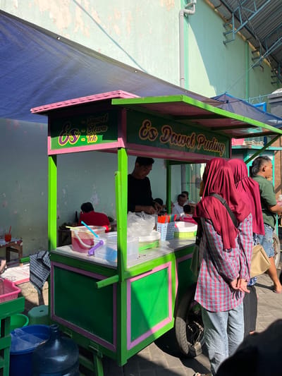 An outdoor, medium shot captures a food cart selling "Es Dawet Puding" under a blue tarp. The cart is painted green with pink trim and features signage indicating its offerings. A vendor, a man in a black shirt, is preparing drinks inside the cart. Two women wearing maroon hijabs and plaid shirts are standing in front of the cart, facing away from the camera. Another man in a green shirt is visible to the right, and several other figures are present in the background. The environment appears to be a street or market area, with a textured, light-colored wall behind the cart and paving stones on the ground. The scene is illuminated by bright sunlight, casting shadows. The cart is equipped with various containers, cups, and straws, suggesting active business. The overall impression is of a bustling daytime scene at a local food stall in Juwet Kenongo, Indonesia.