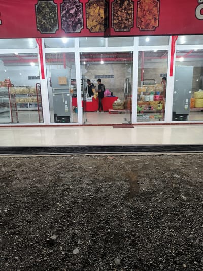The image shows the entrance to a store that sells snacks and chips. The store has a red facade with pictures of various snacks displayed above the entrance. Large glass windows reveal the interior, where shelves are stocked with packaged snacks. Two young men are visible inside the store, appearing to be employees. One is standing behind a counter, and the other is handing a bag of snacks to a customer, whose legs are visible as they are being served. The store's interior appears to be well-lit. A sign with the text "AWAS KACA" (which translates to "Beware of Glass") is posted on a refrigerator inside the store. In front of the store, there is a tiled walkway and an area covered with gravel and small rocks. The lighting suggests it might be evening or a dimly lit indoor environment. The location is identified as Bulo Gogo, Indonesia.