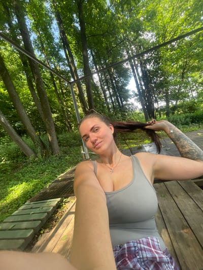 In Мосмек Онт, Russia, a woman is captured in a selfie taken outdoors amidst lush greenery and trees. She is seated on wooden planks, with a rustic wooden table visible. The woman is wearing a gray tank top and a plaid skirt. Her dark hair is partially pulled back, and she is holding a strand of it. A silver cross necklace adorns her neck. Her left arm is adorned with a sleeve tattoo. The background is dominated by the dense foliage of trees and sunlight filtering through the leaves, suggesting a natural, possibly park-like setting.