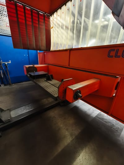 The image shows an industrial setting in Bad Honnef, Germany. In the foreground, there is a metal platform with a perforated surface, likely part of a machine. A black box-like object with a lever is positioned on this platform. To the right, an orange structure extends, with a portion of the brand name "CLO" visible in blue lettering. In the background, red and translucent curtains hang from a ceiling-mounted structure. To the left, a blue wall is visible, with some equipment and metal poles leaning against it. The floor is a dark, concrete surface, with shadows cast by the machinery. The scene appears to be a workshop or factory environment.
