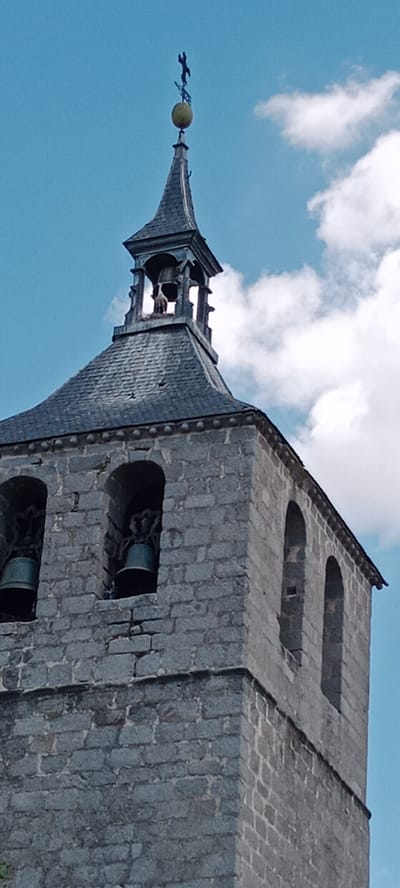The image shows a close-up, low-angle view of a stone bell tower against a blue sky with scattered white clouds. The tower is constructed from rough-hewn grey stones, with some areas showing signs of age and wear. At the top of the tower, there is a dark, steeply pitched roof made of slate tiles, topped by a finial featuring a cross and what appears to be a weather vane. Below the finial, a belfry structure houses at least one bell. Further down the tower, two arched openings are visible, each containing a bell. The bells are dark-colored and suspended from ornate metal frames. The overall impression is of an old, possibly historic, church tower. The context provided indicates that this is the bell tower of the Church of Nuestra Señora de la Asunción in Galapagar, Spain.