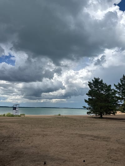 This media file shows a beach scene with dramatic storm clouds gathering. The foreground is dominated by a sandy beach, with dry grass and a single tree providing a touch of greenery. In the background, a calm body of water stretches to the horizon, with a faint treeline visible in the distance. The sky is filled with a mix of dark, heavy storm clouds and brighter, sunlit clouds, suggesting an impending change in weather. The time of day appears to be daytime, likely afternoon, given the lighting.

There is a small, dark-colored structure with a sign on it, located near the water's edge. The sign appears to have some text and graphics, but it is too small and blurry to be legible. There are also a few people faintly visible on the beach, appearing as small figures in the distance. The overall mood of the image is one of natural beauty and a sense of anticipation due to the approaching storm. The location is identified as Володарский ауылдық округі, Kazakhstan, which is consistent with a beach or lakeside environment.