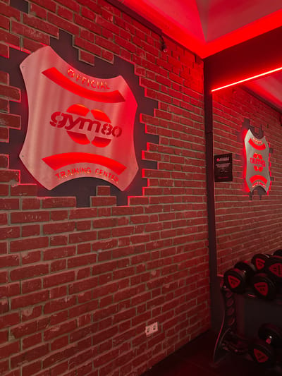 The image depicts the interior of a gym with a brick wall background illuminated by red lighting. A metal shield-shaped logo with the text "OFFICIAL GYM80 TRAINING CENTER" is prominently displayed on the left side of the wall. To the right, another similar logo with the text "GYM80" is visible, along with a small sign with German text and a rack of dumbbells. The overall aesthetic suggests a modern and edgy gym environment. The specific location in Wuppertal, Germany is not discernible from the image alone.