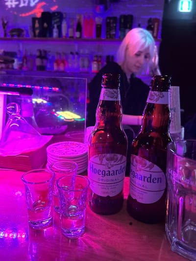 The image depicts an indoor scene within a bar or pub located in 마포구, South Korea. The environment is dimly lit with prominent purple and pink neon lighting.
In the foreground, a wooden bar counter displays two full or nearly full bottles of Hoegaarden Original beer, three empty clear shot glasses, and one larger empty clear drinking glass. To the left of the beer bottles, a stack of white circular coasters is visible, next to a stack of square paper napkins under a clear glass carafe or pitcher. A reflective surface, possibly a mirror or glass partition, stands behind these items, showing reflections of multicolored neon lights.
In the mid-ground and background, behind the bar counter, a person with short, light-colored hair, likely platinum blonde or white, is visible. This individual, presumably a bartender, is wearing dark clothing and is positioned with their head tilted downwards, appearing to be focused on an activity behind the bar. Behind the bartender, multiple shelves stocked with various bottles of alcohol are illuminated by ambient lighting. The overall scene suggests an active evening service at a contemporary establishment.