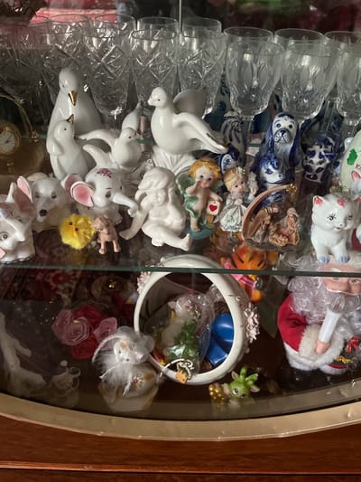This media file displays a close-up, top-down view of a glass display cabinet filled with various decorative figurines. The subjects are primarily ceramic figurines, including several white doves, a sleeping baby, and a collection of rabbit figurines with pink accents. There are also other small trinkets such as a small green dragon-like creature, a yellow fuzzy chick, and what appears to be a small clock. In the background, crystal glasses are visible, stacked in rows. The overall scene is a collection of decorative items, likely for display rather than any active event. There are no visible location cues or text. The lighting appears to be artificial, suggesting the display is indoors. The style of the figurines suggests a whimsical and perhaps sentimental collection.
