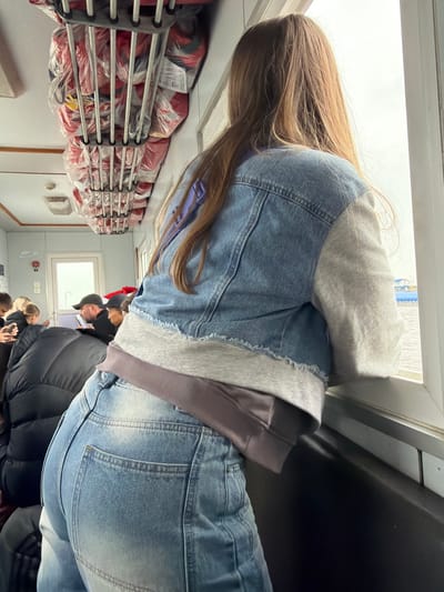 The image shows the interior of what appears to be a ferry or passenger boat, seen from a slightly elevated, side-angle perspective. The primary subject in the foreground is a person with long, straight brown hair, facing away from the camera and looking out a large window. They are wearing a light blue denim jacket over a grey hooded sweatshirt, with the sleeves of a dark purple or grey undershirt visible, and light blue denim jeans.

Along the left side of the vessel's interior, multiple red life jackets, each individually sealed in clear plastic bags, are neatly stored on grey metal racks. The racks extend along the wall, indicating a safety-conscious environment. In the mid-ground and background, other passengers are visible, mostly from behind or the side. They are dressed in winter attire, including puffer jackets and hats, suggesting cool weather. Some appear to be looking out of the windows or standing in a queue.

Through the large windows, a bright, overcast sky is visible, suggesting daytime. The overall lighting is bright and even, consistent with natural light from large windows on a cloudy day. The setting implies travel or a journey. Given the location context of Nizhneleninskoe, Russia, this is likely a ferry crossing the Amur River, which connects Russia and China. No discernable text is clearly visible on any objects or surfaces.