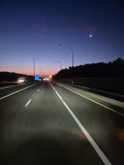 The image depicts a highway scene at dusk or dawn, likely on the E19 motorway near Schoten, Belgium. The sky is a deep twilight blue, with a sliver of a crescent moon visible in the upper right corner. The road is asphalt, with clear white lane markings indicating multiple lanes in each direction. Streaks of light suggest motion blur from the perspective of a vehicle traveling at speed.
On the left side of the highway, there are trees lining the road, with some appearing as dark silhouettes against the fading light. Streetlights with long poles are spaced along the highway, casting illumination onto the road surface. In the distance, there are visible headlights from other vehicles, suggesting moderate traffic. A blue road sign with white lettering is present on the left, indicating directions and destinations, though the text is too small to read clearly. On the right side of the highway, a darker, textured barrier or sound wall is visible, also lined by trees. The overall impression is of a smooth, well-maintained highway during a transitional time of day, with the artificial lights beginning to dominate the natural light. There are no visible people.