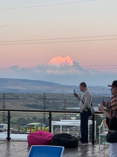 Two men are standing on a wooden deck, looking out at a landscape with mountains and a town below. The sky is a mix of blue and pink, suggesting it is either sunrise or sunset. One man, wearing a plaid shirt and a baseball cap, is holding a phone and a cup. The other man, wearing a light-colored sweatshirt, is also holding a phone and is bending over slightly. In the background, there are power lines and what appears to be a large, puffy cloud formation that is lit up with an orange glow. On the deck, there are also some outdoor furniture, including a pink beanbag chair and a blue chair. The overall mood seems relaxed and contemplative, with the men engrossed in their phones while taking in the view. The location is identified as Mukhatskaro, Georgia. There is a license plate visible on a lime green car in the background, reading "T711JS".