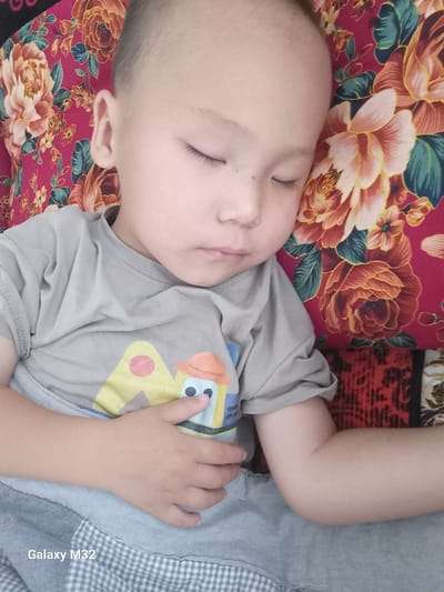 A young child, appearing to be asleep, is lying on their back. The child has short dark hair and is wearing a light gray t-shirt with a graphic design. The child's head rests on a pillow with a vibrant floral pattern featuring large red and orange flowers against a dark pink background. The child's left arm is bent at the elbow and rests across their chest. The background behind the pillow is a patterned fabric in shades of red and black, possibly part of a bedspread or furniture. The image is taken from a high angle, looking down at the child. The location within Ынтымак, Kyrgyzstan, is not determinable from the visual information provided in the image. The bottom left corner of the image contains the text "Galaxy M32".