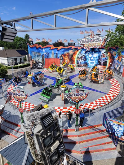 This image depicts the "Ghost Rider" amusement park ride in the city of Best, Netherlands. The ride, a spinning amusement ride with individual vehicles resembling motorcycles, is centrally located on a checkered red and white circular platform. The backdrop of the ride features a large mural with a prominent skull, flames, and cityscapes, along with American flags. Several people are visible on and around the ride, some seated in the vehicles and others standing on the surrounding platforms. The surrounding environment includes trees, a building with a tiled roof, and blue railings. The lighting suggests it is a bright, sunny day.
