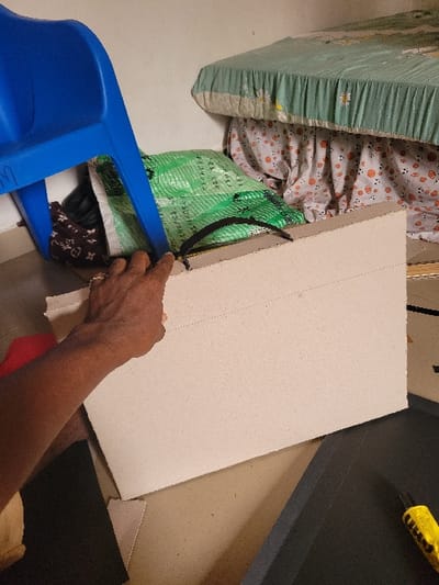 The scene is an indoor room in Puerto López, Colombia. In the foreground, a dark-skinned hand, wearing a ring on the fourth finger, grips a black fabric handle attached to a large, light-brown cardboard panel. The cardboard panel features perforated lines across its top surface. The floor beneath the cardboard appears light-colored. To the lower right, a dark gray foam-like material and a yellow object, resembling a utility knife, are visible on the floor. In the background-left, an inverted blue plastic chair rests above a green woven plastic bag and a dark bag with a light-colored monogram-like pattern. Behind these items, a bed is visible, covered by a light green quilted bedspread with a white floral pattern. A white sheet with an orange polka-dot pattern drapes from beneath the bedspread. A plain white wall forms the background.