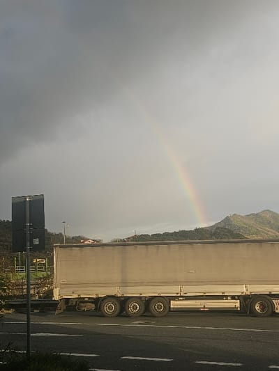 The image displays a roadside scene in Sestri Levante, Italy, characterized by a large vehicle and atmospheric conditions. A light-colored semi-trailer truck occupies the midground, positioned horizontally on a paved road. The truck's trailer features multiple axles with dark wheels and a corrugated side facing the viewer. The road in the immediate foreground exhibits white dashed and solid lane markings. To the far left, a dark vertical pole supports two rectangular, non-descript signs.
The background consists of a mountainous landscape under a mixed sky. On the left side, green, forested hills are visible, dotted with scattered residential structures. To the right, a prominent, rugged mountain peak rises, partially illuminated by sunlight. The sky transitions from a dense, dark overcast on the left to a lighter, brighter grey on the right. A distinct, colorful rainbow arc is visible in the upper right quadrant of the sky, appearing to descend towards the illuminated mountain peak. No individuals are present in the frame, and the truck appears to be stationary or in transit.