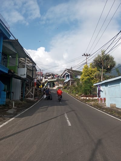The image shows a street scene in Mya Sit Kaing Ywar Thit, Myanmar. Two motorcycles are moving away from the viewer on an asphalt road with a white line marking the center. The motorcycle in the lead has a rider wearing a red shirt, while the second motorcycle carries two people and what appears to be a large black tire.
The road is lined with buildings of various designs and colors, mostly with corrugated metal roofs. Some appear to be residences, while others may be shops or businesses. Electrical wires are strung overhead. Vegetation, including trees and smaller plants, grows along the roadside and around the buildings. The sky is partly cloudy with a mix of blue sky and white clouds. A person is standing on some scaffolding beside a building in the mid-ground.
